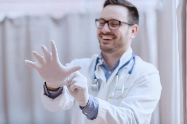 身穿白衣 面带微笑的年轻白人男医生 脖子上戴着眼镜和听诊器 在检查病人之前戴上橡胶手套 有选择地把重点放在手头上 — 图库照片