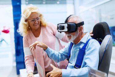 Smiling bearded Caucasian man sitting in the chair and truing out virtual reality goggles while his wife standing next to him and smiling. Tech store interior. clipart