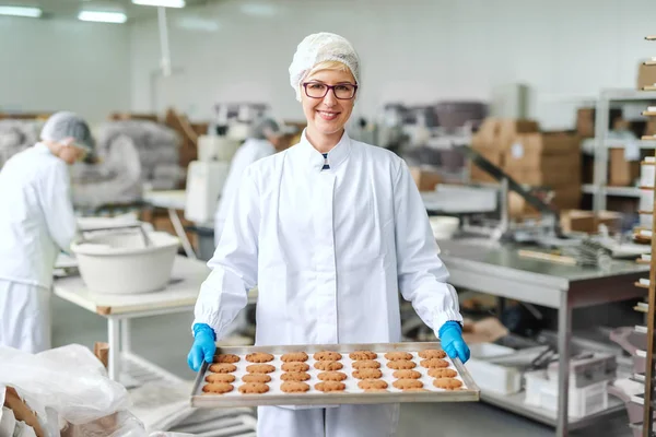 の笑顔金髪白人の従業員は 無菌の制服で 眼鏡の立って クッキーとトレイを保持します 食品工場内装 バックグラウンドで他の従業員が働く — ストック写真