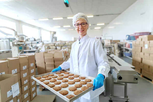 Smiling blonde Caucasian employee in sterile uniform and with eyeglasses standing and holding tray with cookies. Food factory interior. In background other employees working.