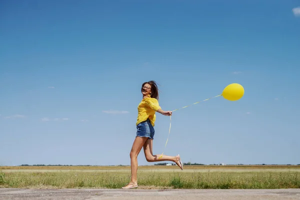 华丽的白种少女在黄色上衣 牛仔短裤和眼镜运行户外与黄色气球在手 — 图库照片