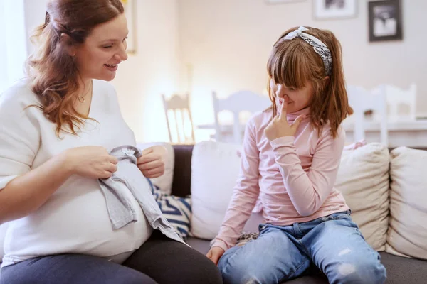 快乐怀孕的白种人妈妈坐在她可爱的女儿在客厅的房间里 抱着婴儿的衣服 — 图库照片
