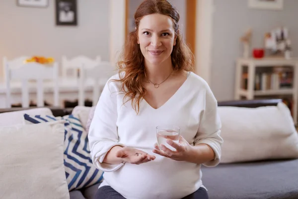 片手にビタミンを持ち もう一方の手に水のグラスを持つ笑顔の白人の妊婦 — ストック写真