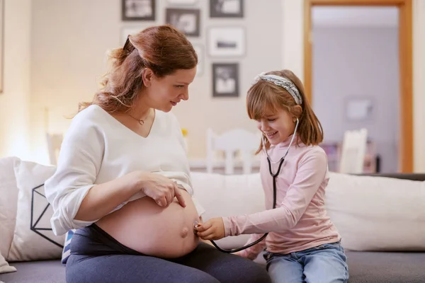 美丽的白种人怀孕的母亲坐在客厅与她的女儿 女儿听听听器婴儿的心跳 客厅内部 — 图库照片