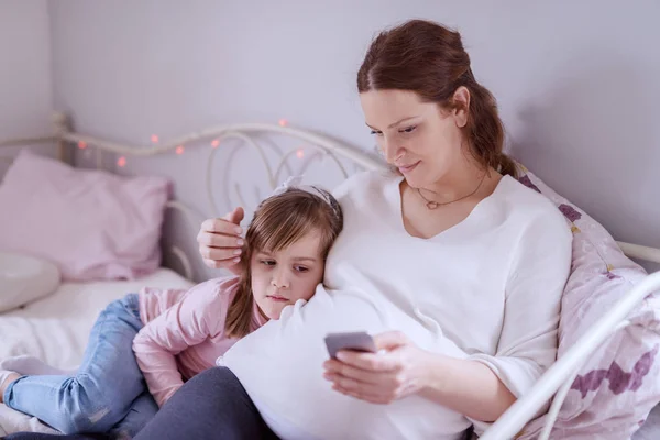 美丽的白种孕妇坐在坐在一个 拥抱她的女儿 拿着智能手机 — 图库照片