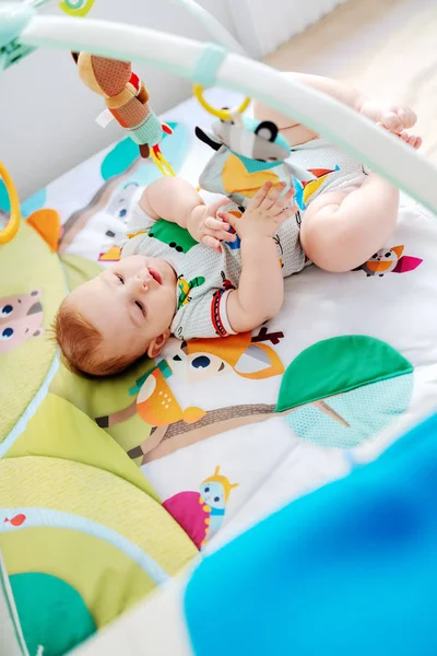 Top view of playful Caucasian chubby six months old baby boy in colorful bodysuit playing with crib toys.