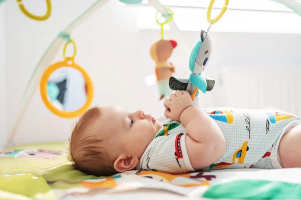 Side view of playful Caucasian chubby six months old baby boy in colorful bodysuit playing with crib toys.