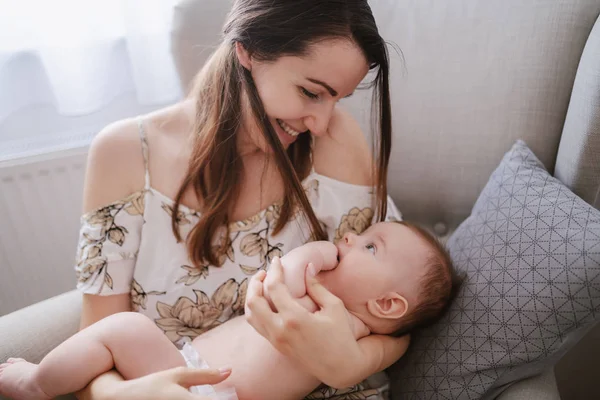 若い白人の母親が アームチェアに座りながら6ヶ月の息子を抱きしめて世話をする 赤ちゃんは目に母親を見て 口に指を入れて — ストック写真