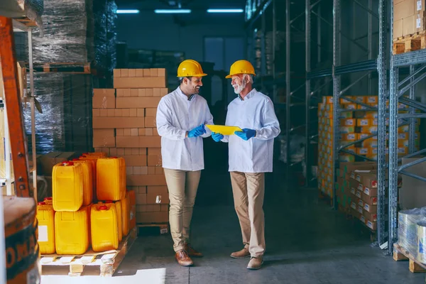 两名面带微笑的仓库工人身着白色制服 头戴黄色头盔 站在一起谈论工作 较旧的一个持有文件夹 手拿文档 — 图库照片