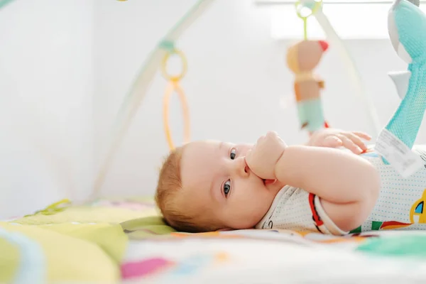 아름다운 개월된 아이가 장난감에 둘러싸여 침대에 카메라를 엄지손가락을 — 스톡 사진