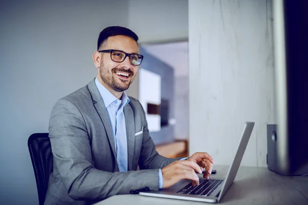 Smiling attractive Caucasian white collar worker with eyeglasses sitting at office, looking at colleague and using laptop.