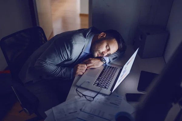 工作过度的专职员工穿着灰色西服 晚上睡在办公室里 桌上放着报告 眼镜和笔记本电脑 — 图库照片