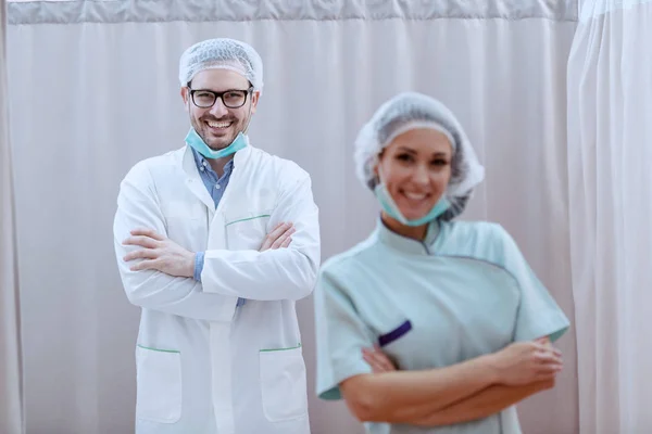 身穿制服 戴着面具 头戴发网 双臂交叉站在诊所的友善的年轻白人医生的画像 在前景护士摆姿势 对医生的选择性关注 — 图库照片