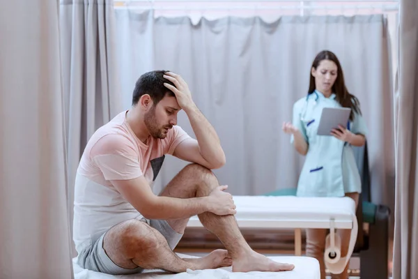 年轻的白人男性病人坐在病床上 感到疼痛 背景女性护士使用平板电脑 — 图库照片