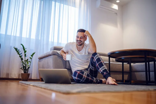 Charismatic smiling caucasian man in pajamas sitting on the floor in living room, leaning on sofa, holding mug with coffee and looking at laptop. Weekend activities concept.