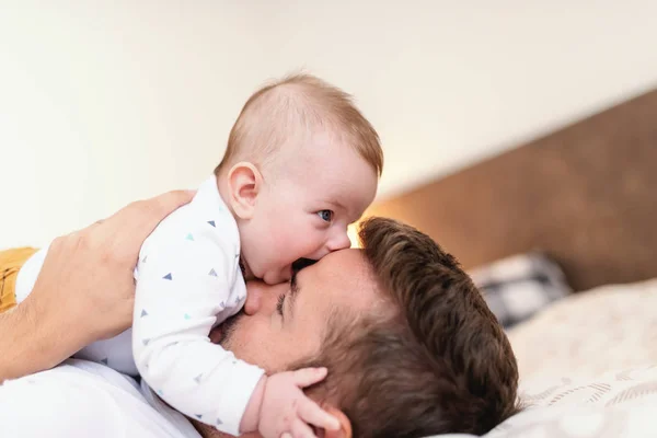 幸せな白人のハンサムなお父さんは寝室でベッドに横たわって 彼の胸に彼の愛らしい6ヶ月の息子を保持しています 赤ちゃんがパパの顔を噛む無条件愛の概念 — ストック写真