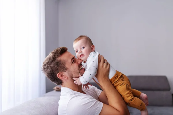 遊び心のあるハンサムな白人の若いお父さんは リビングルームでソファに座っている間 彼の愛する6ヶ月の男の子を持ち上げる 赤ちゃんは父との時間を楽しんで カメラを見て — ストック写真