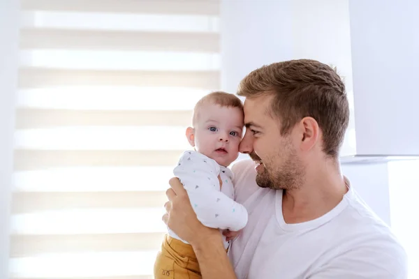 愛らしい愛する息子を抱きしめ 窓の横に立ちながら抱き合うハンサムな白人の若いお父さんを誇りに思う 赤ちゃんはカメラを見ている — ストック写真