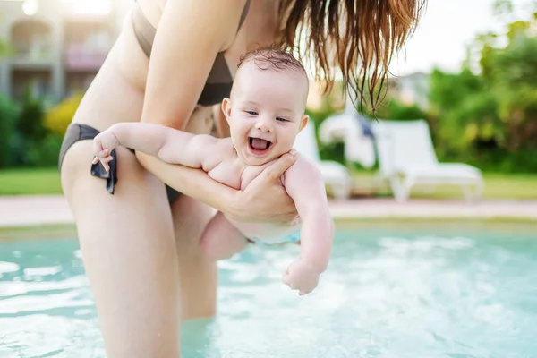 Cheerful Caucasian 6 months old baby boy learning how to swim at swimming pool. Mother holding her son. First time at pool concept.