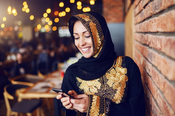 美丽的微笑的穆斯林妇女穿着传统服装坐在咖啡店 并使用智能手机发送或阅读信息的肖像 — 图库照片