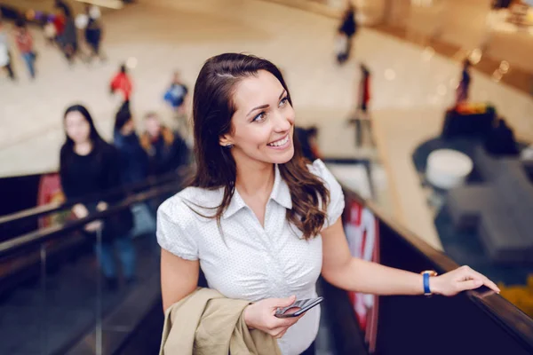 Menakjubkan Tersenyum Brunette Memanjat Eskalator Memegang Jaket Krem Dan Ponsel — Stok Foto