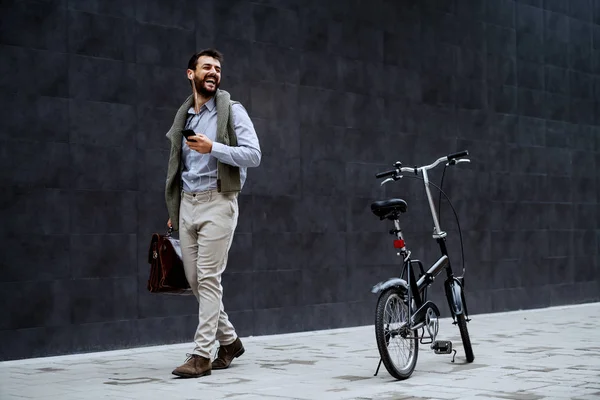 微笑着英俊时髦的高加索商人一边听音乐 一边拿着公文包去工作 他旁边是自行车 背景是灰墙 — 图库照片