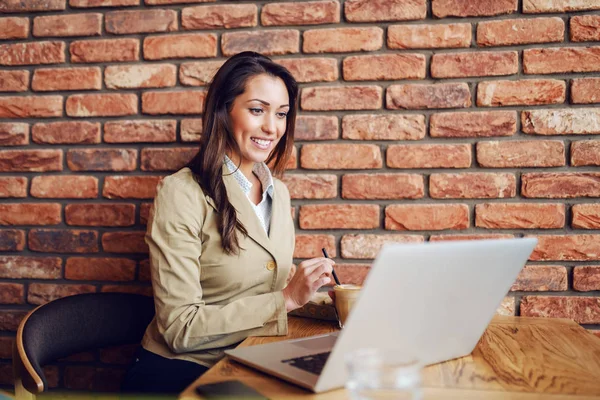 专注的微笑女商人穿着智能休闲坐在咖啡馆 搅拌咖啡 看着笔记本电脑 — 图库照片