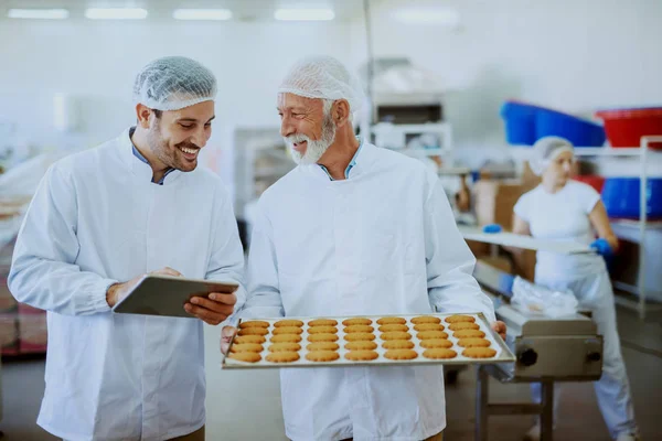 食品工場でクッキーとトレイに立って滅菌白の均一性の高い大人の従業員 彼の隣に立って監督者 錠剤を保持し 食品の品質をチェック — ストック写真