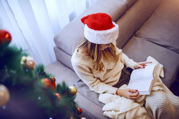 穿着毛衣 头戴圣诞礼帽 头戴圣诞礼帽 坐在圣诞树旁沙发上看书的美丽的高加索女青年的头像 — 图库照片