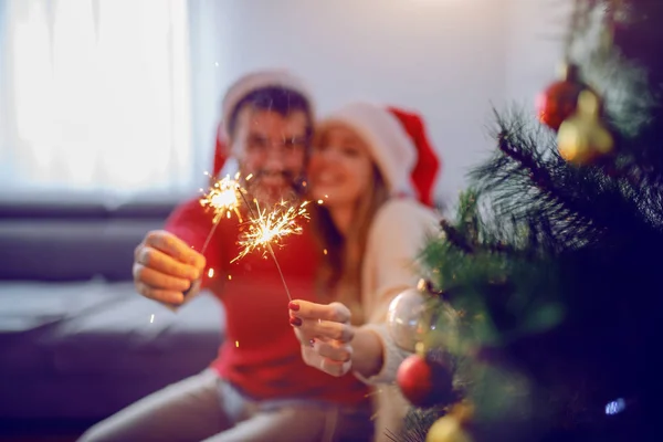 快乐的高加索夫妇 头上戴着圣诞帽 坐在沙发上 拿着洒水器 庆祝新年 前景光明的是圣诞树 有选择地注重手部 — 图库照片