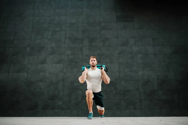 Front view of handsome muscular bearded caucasian man in shorts and t-shirt holding training bag while doing lunges.