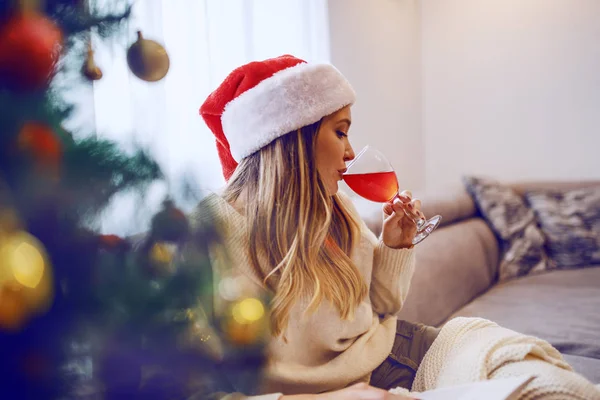 リビングルームのソファに座って ワインを飲んで頭の上にサンタの帽子を持つ豪華な白人ブロンドの女性の側面図 手前はクリスマスツリー — ストック写真