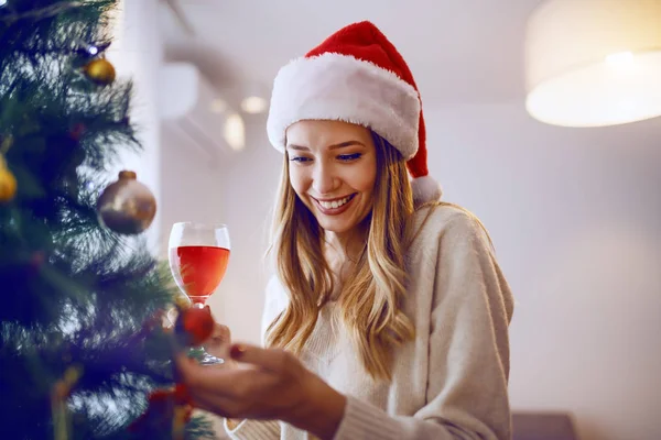 豪華な笑顔Caucasianブロンド女性とともにサンタ帽子でタッチオーナメント上のクリスマスツリーと飲酒ワインながらリビングルームに立って — ストック写真