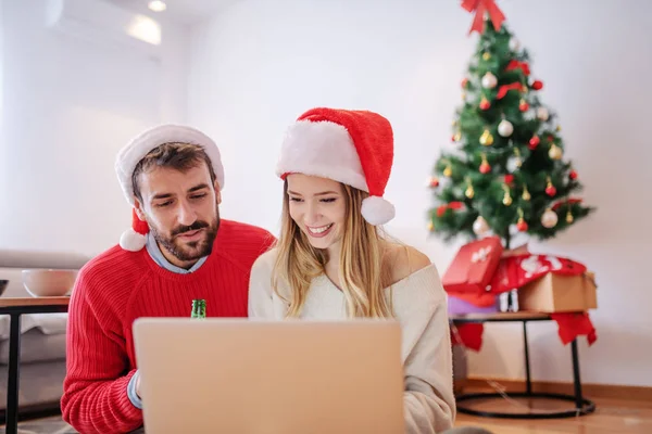 幸せなハンサムな白人のカップルは 頭の上にサンタの帽子とラップトップを使用してリビングルームに座っています 背景にはプレゼント付きクリスマスツリーがあります — ストック写真