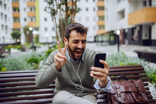 웃으며 잘생긴 사업가가 스마트폰으로 비디오 통화를 공원에 벤치에 승리의 표시를 — 스톡 사진