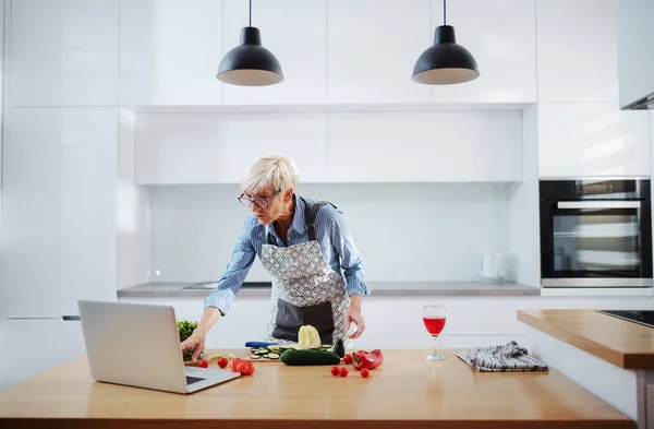 短い髪とエプロンで見栄えの良い白人のシニア女性は ラップトップ上のレシピに従って キッチンに立っている間に食事を準備します キッチンカウンターでは ノートパソコン 野菜やワインのガラスです — ストック写真