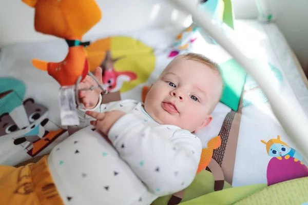 Curious smiling baby boy six months old dressed in bodysuit lying in bed and playing with his crib toys while looking at camera.