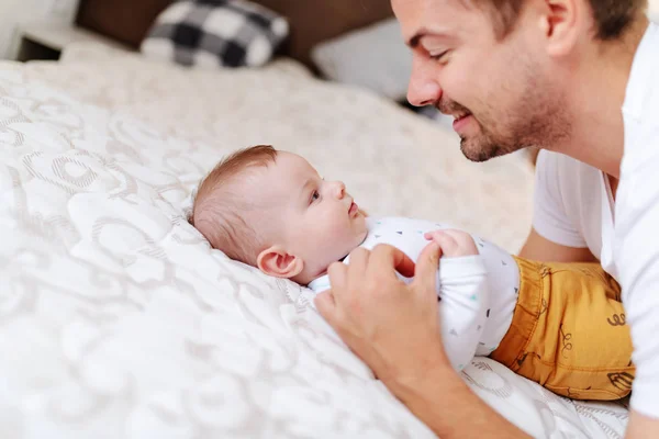 ハンサムな若いお父さんの側のビューを再生し 彼の愛らしい6ヶ月の息子と話をする ベッドに寝そべってる間に父親を見て — ストック写真