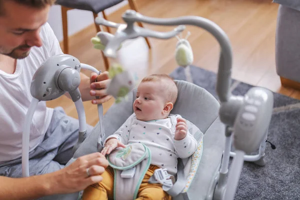 Handsome smiling young caucasian dad putting his adorable 6 months old son in baby rocker chair. Baby looking confused at father.