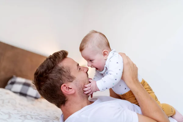 ハンサムな若いお父さんは寝室でベッドに横たわって 彼の愛する笑いの男の子6ヶ月を持ち上げる 無条件愛の概念 — ストック写真