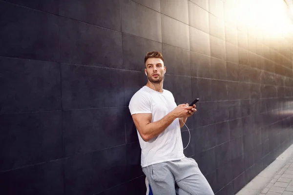 Beautiful caucasian bearded blonde muscular man in t-shirt holding smart phone while leaning on the wall and looking away. In ears are earphones.