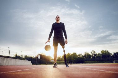 Full length of sporty caucasian handicapped man in sportswear and artificial leg dribble the ball while standing on racetrack. clipart
