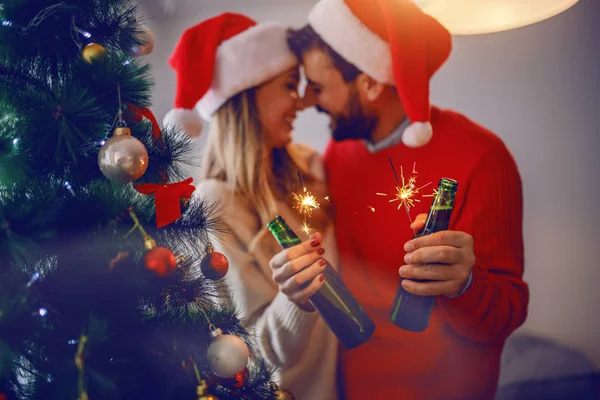 リビングやスヌグリングに立ちながらビールやスパークリングを持って頭の上にサンタの帽子と喜びの白人カップル 手前はクリスマスツリー 手に焦点を当てる — ストック写真