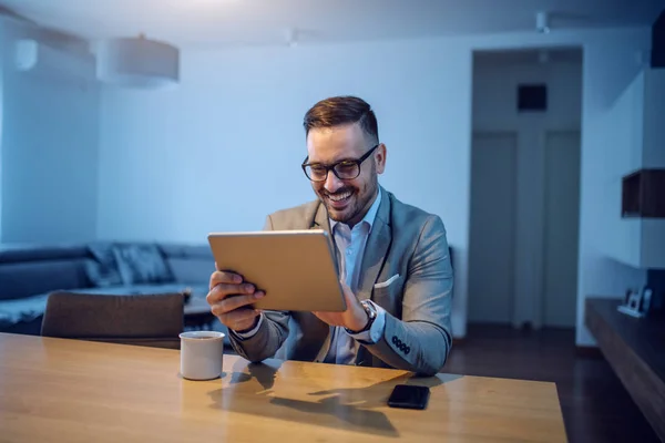 幸せな上品な白人ハンサムなビジネスマンスーツと眼鏡ダイニングテーブルに座って タブレットを使用しています テーブルの上には朝のコーヒーとスマートフォンがあります 背景にリビングルームがあります — ストック写真