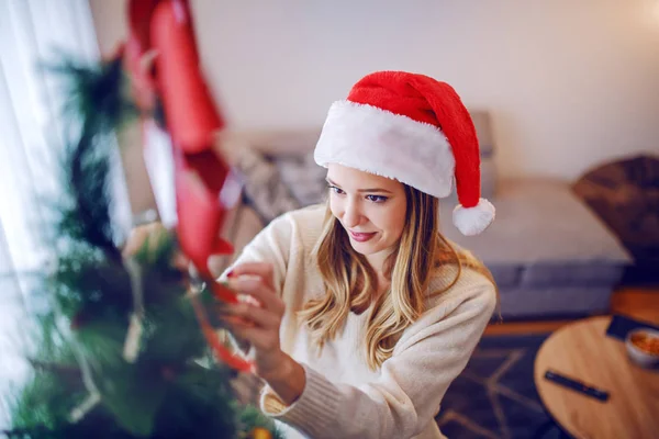 身穿毛衣 头戴圣诞帽 装饰圣诞树 站在客厅里的美丽的高加索女青年 — 图库照片