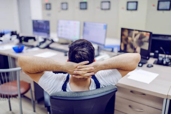 Rear view of lazy factory worker sitting on chair with hands behind head and sleeping in control room.