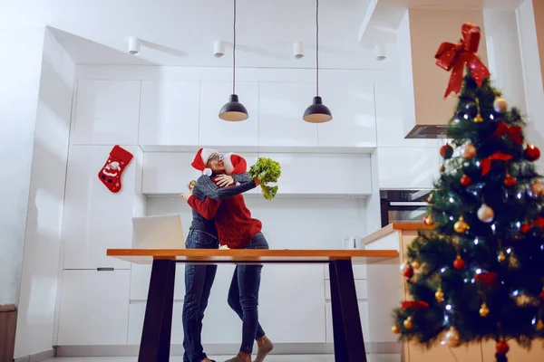 怀孕的女儿站在厨房里抱着妈妈 两个头上都戴着桑塔帽 老女人拿着莴苣 厨房柜台上有笔记本电脑 在前景中的是圣诞树 — 图库照片