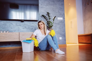 Tired worthy caucasian blonde housewife sitting on the floor in living room and looking at bucket with cloth and sponge on it. clipart