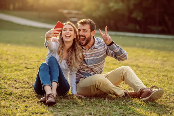 かわいい幸せな笑顔白人のカップルは 自然の中で牧草地に座って自撮りを取る 男巨大な女性と勝利の兆候を示しながら 女性はスマートフォンを保持します — ストック写真