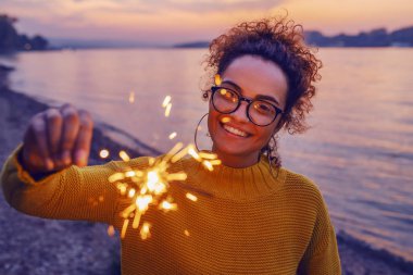 Cute smiling mixed race hipster girl with eyeglasses and curly hair standing on shore next to river and holding sparkler. clipart
